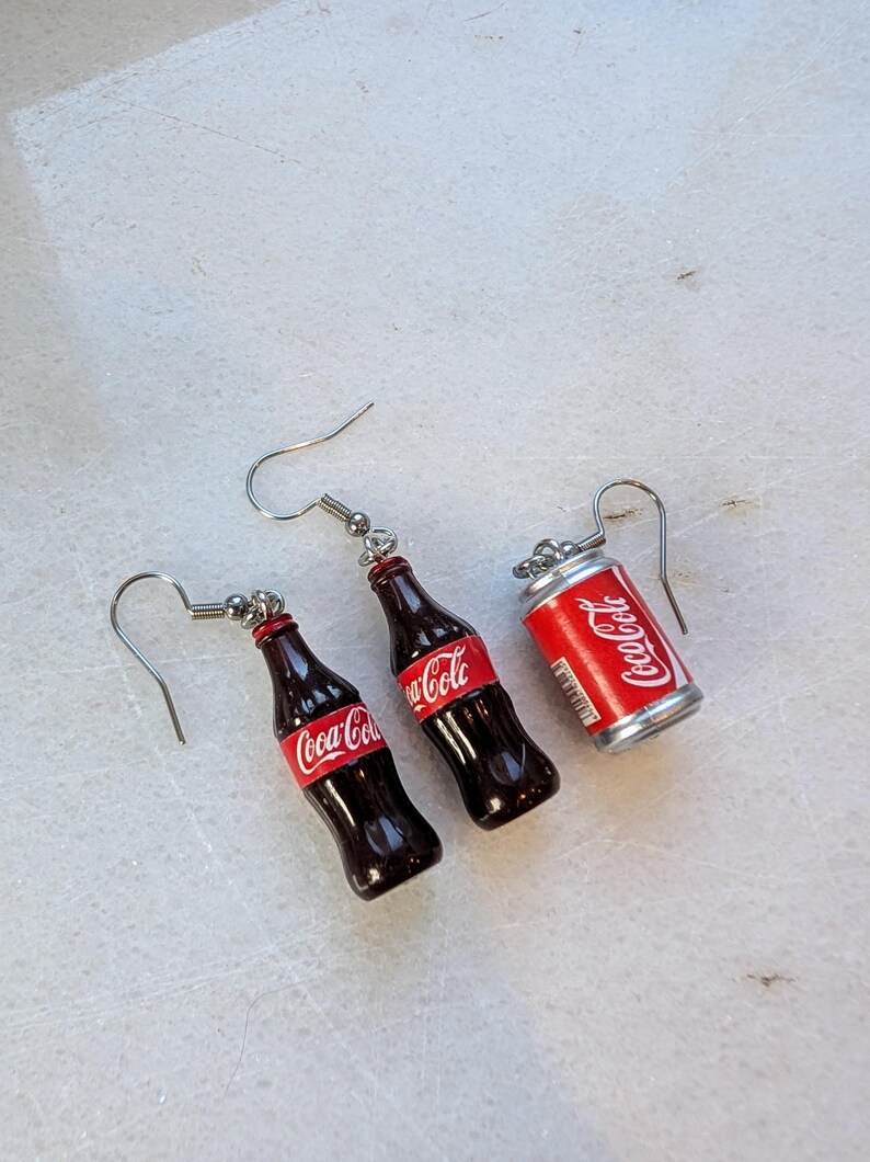 Puede incluir: Un par de pendientes colgantes de plata con botellas de Coca-Cola en miniatura y una lata. Las botellas son de vidrio marr&oacute;n con etiquetas de Coca-Cola rojas y blancas. La lata es de plata con una etiqueta de Coca-Cola roja y blanca.