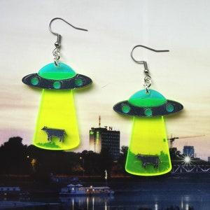 May include: A pair of neon green and blue UFO earrings with a black silhouette of a cow inside each UFO. The earrings have silver hooks.