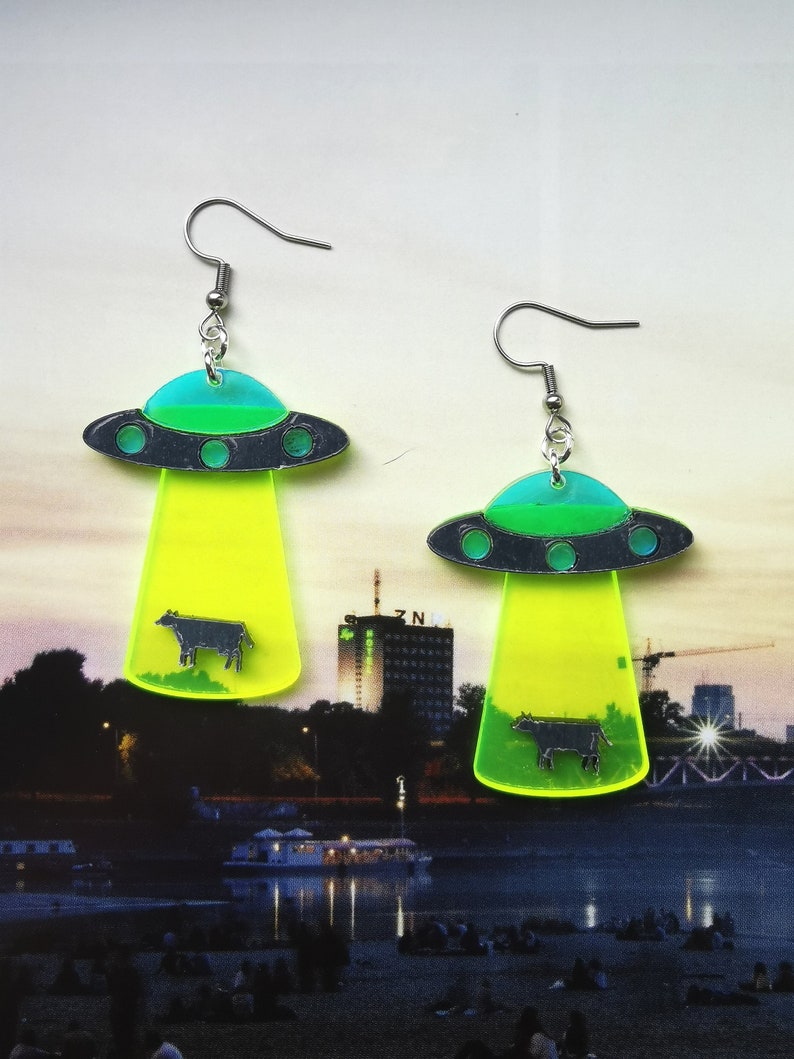 May include: A pair of neon green and blue UFO earrings with a black silhouette of a cow inside each UFO. The earrings have silver hooks.