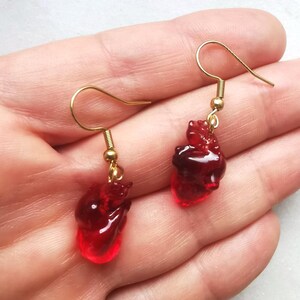 May include: A pair of gold dangle earrings with red heart-shaped charms. The hearts are translucent and have a slightly textured surface.