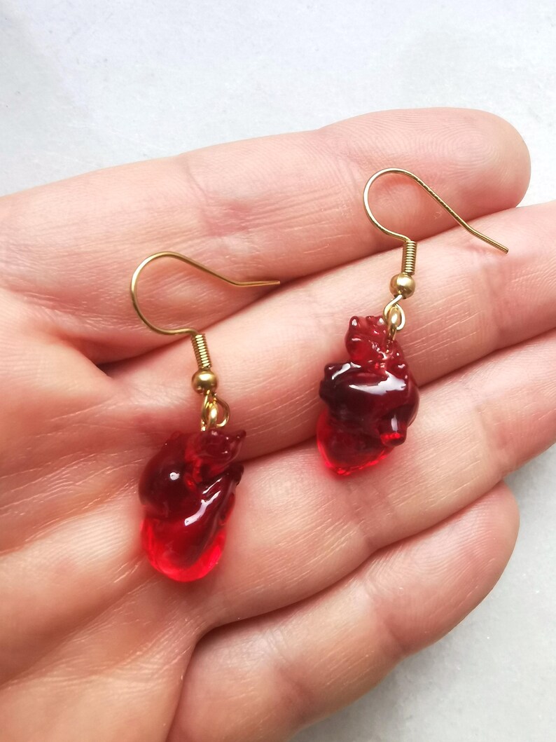 May include: A pair of gold dangle earrings with red heart-shaped charms. The hearts are translucent and have a slightly textured surface.