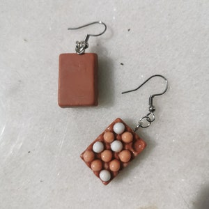 Peut inclure: Une paire de boucles d'oreilles en forme de carton d'&oelig;ufs brun et blanc. Les boucles d'oreilles sont en p&acirc;te polym&egrave;re et ont un design r&eacute;aliste de carton d'&oelig;ufs. Le carton d'&oelig;ufs est rempli d'&oelig;ufs blancs et bruns.