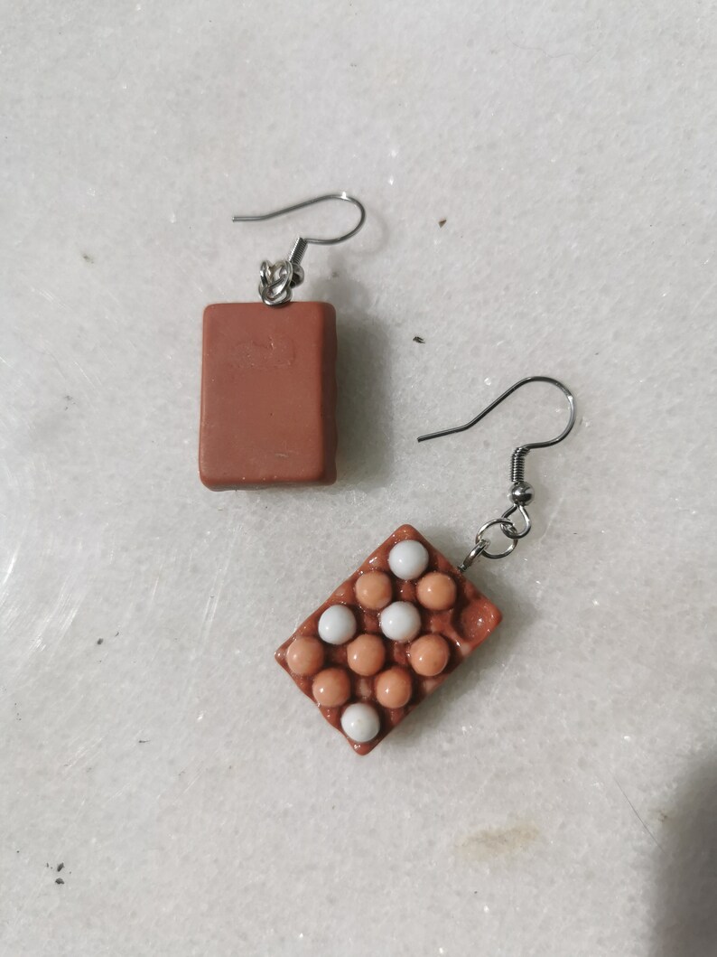Peut inclure: Une paire de boucles d'oreilles en forme de carton d'&oelig;ufs brun et blanc. Les boucles d'oreilles sont en p&acirc;te polym&egrave;re et ont un design r&eacute;aliste de carton d'&oelig;ufs. Le carton d'&oelig;ufs est rempli d'&oelig;ufs blancs et bruns.