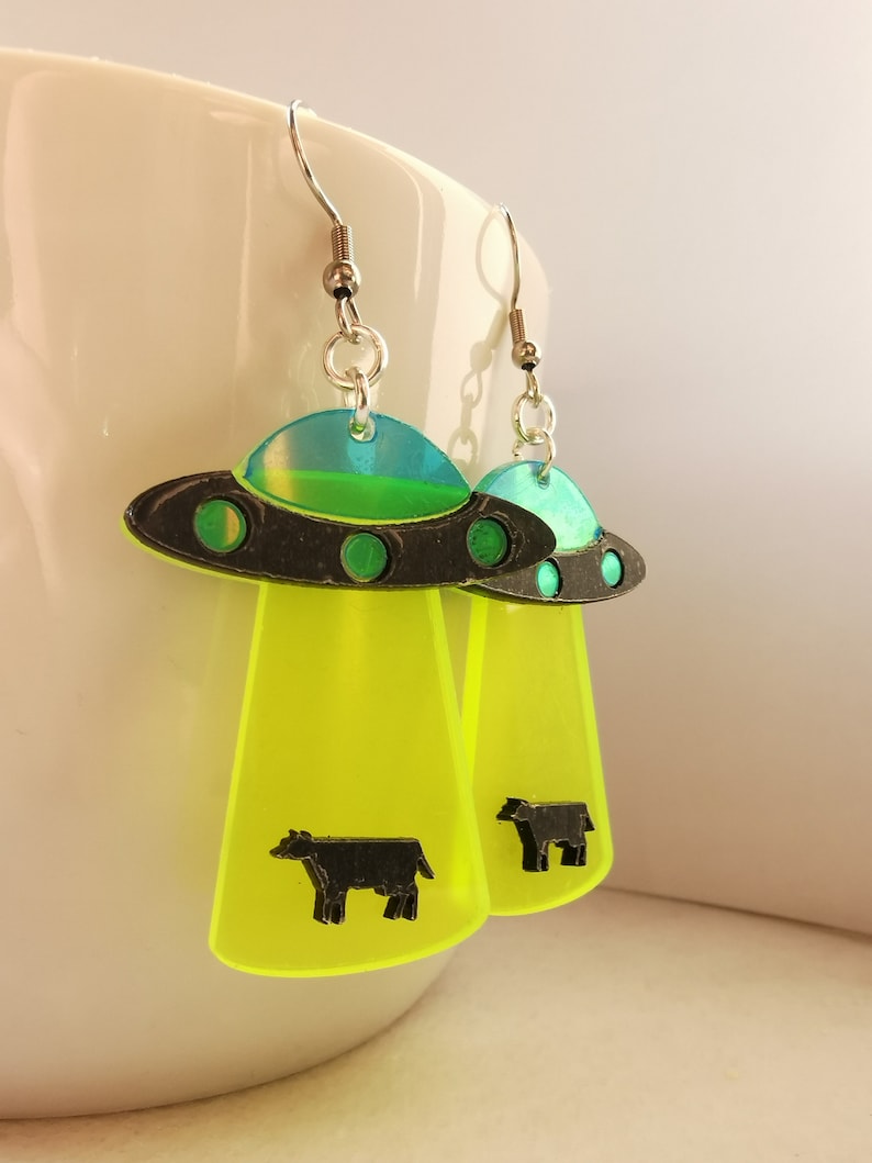 May include: A pair of neon green and black UFO earrings with a black cow silhouette on each earring.