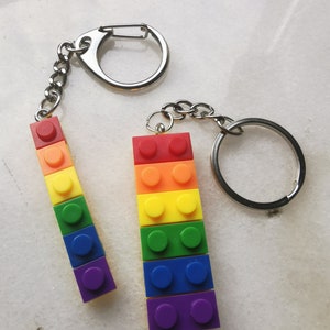 May include: Two rainbow colored keychain charms shaped like Lego bricks. The charms are attached to silver key rings.