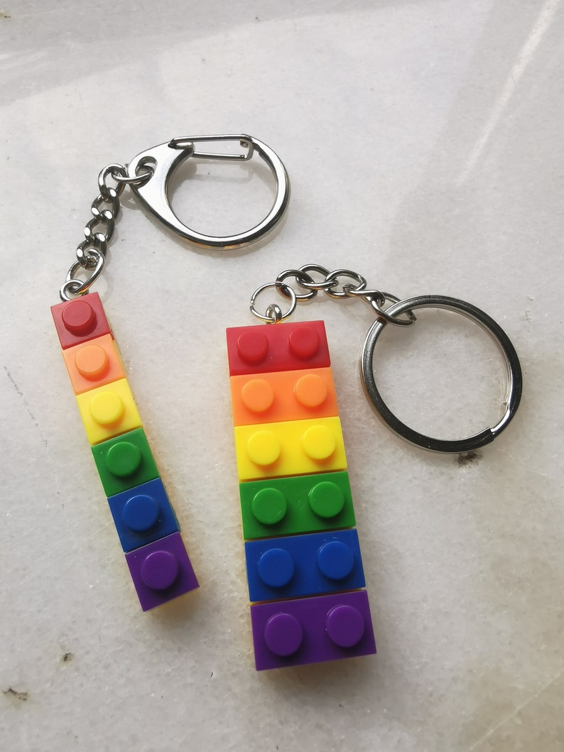 May include: Two rainbow colored keychain charms shaped like Lego bricks. The charms are attached to silver key rings.