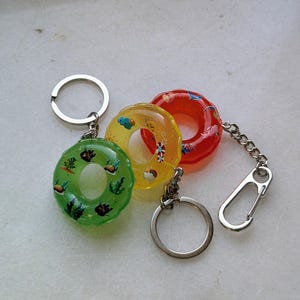Puede incluir: Tres llaveros en miniatura con forma de salvavidas, de colores verde, amarillo y rojo. El verde tiene motivos marinos, el amarillo nubes y el rojo conchas. Cada uno tiene un llavero y una cadena plateados.