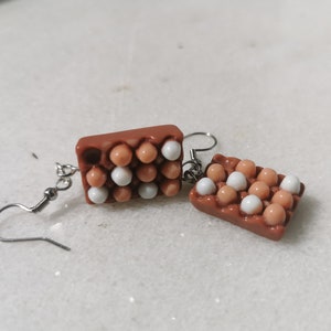 Peut inclure: Une paire de boucles d'oreilles en forme de bo&icirc;tes &agrave; &oelig;ufs avec des &oelig;ufs blancs et bruns. Les boucles d'oreilles sont en p&acirc;te polym&egrave;re et ont des crochets argent&eacute;s.