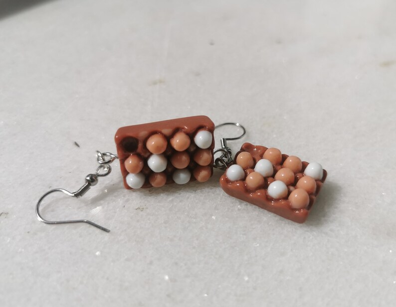 Peut inclure: Une paire de boucles d'oreilles en forme de bo&icirc;tes &agrave; &oelig;ufs avec des &oelig;ufs blancs et bruns. Les boucles d'oreilles sont en p&acirc;te polym&egrave;re et ont des crochets argent&eacute;s.