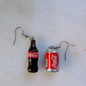 Puede incluir: Un par de pendientes de aro de plata con encantos en miniatura de botella y lata de Coca-Cola. La botella es marr&oacute;n con un logotipo de Coca-Cola rojo. La lata es plateada con un logotipo de Coca-Cola rojo.