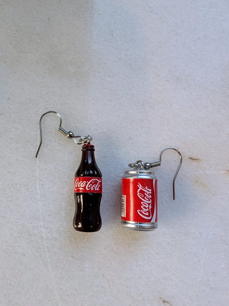 Puede incluir: Un par de pendientes de aro de plata con encantos en miniatura de botella y lata de Coca-Cola. La botella es marr&oacute;n con un logotipo de Coca-Cola rojo. La lata es plateada con un logotipo de Coca-Cola rojo.