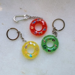 Puede incluir: Tres llaveros en miniatura con forma de salvavidas, en rojo, amarillo y verde. El rojo tiene diseños acuáticos, el amarillo tiene diseños de sol y flores, y el verde tiene diseños de hojas. Cada llavero tiene una cadena y un anillo plateados.