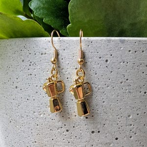 May include: A pair of gold-colored earrings featuring miniature coffee maker charms. The earrings have a simple hook design and are suspended from a small gold bead. The coffee maker charms have a detailed, three-dimensional design.
