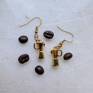 May include: Gold-colored coffee pot earrings with gold-colored ear wires. The earrings are next to several dark brown coffee beans, scattered on a white surface. The earrings are a miniature version of a stovetop espresso maker.