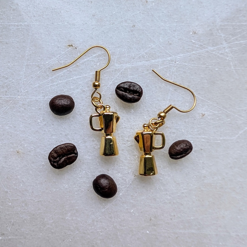 May include: Gold-colored coffee pot earrings with gold-colored ear wires. The earrings are next to several dark brown coffee beans, scattered on a white surface. The earrings are a miniature version of a stovetop espresso maker.