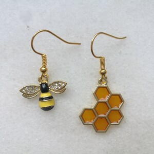 K&ouml;nnte beinhalten: Ein Paar goldene Ohrringe mit einem Bienen- und Wabenmuster. Der Bienenohrring ist gelb und schwarz mit klaren Strasssteinen. Der Wabenohrring ist golden mit einem gelben Wabenmuster.