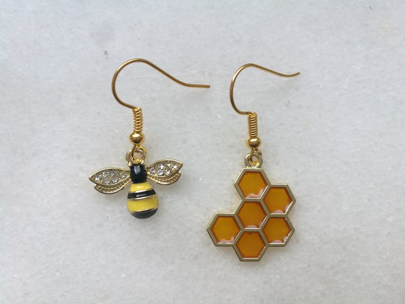 K&ouml;nnte beinhalten: Ein Paar goldene Ohrringe mit einem Bienen- und Wabenmuster. Der Bienenohrring ist gelb und schwarz mit klaren Strasssteinen. Der Wabenohrring ist golden mit einem gelben Wabenmuster.