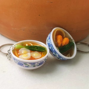 May include: Two miniature ceramic bowls with blue and white floral designs, filled with a simulated noodle soup with vegetables and dumplings. The bowls are attached to keychains.