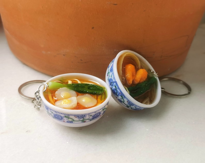May include: Two miniature ceramic bowls with blue and white floral designs, filled with a simulated noodle soup with vegetables and dumplings. The bowls are attached to keychains.