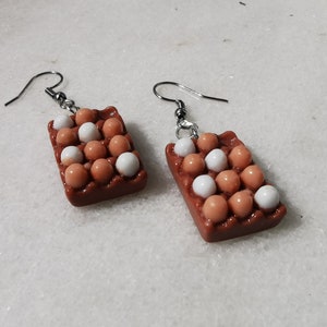 Peut inclure: Une paire de boucles d'oreilles en forme de bo&icirc;tes &agrave; &oelig;ufs remplies d'&oelig;ufs blancs et bruns. Les boucles d'oreilles sont en p&acirc;te polym&egrave;re brune et blanche et ont des crochets argent&eacute;s.