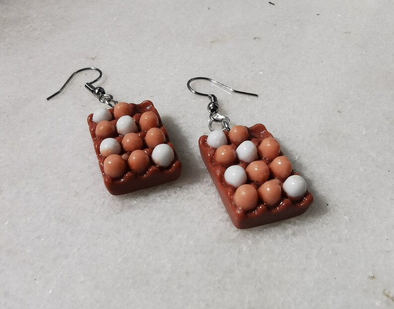 Peut inclure: Une paire de boucles d'oreilles en forme de bo&icirc;tes &agrave; &oelig;ufs remplies d'&oelig;ufs blancs et bruns. Les boucles d'oreilles sont en p&acirc;te polym&egrave;re brune et blanche et ont des crochets argent&eacute;s.