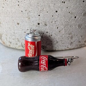 Puede incluir: Un par de pendientes con tem&aacute;tica de Coca-Cola. Un pendiente es una lata de Coca-Cola en miniatura y el otro es una botella de Coca-Cola en miniatura.