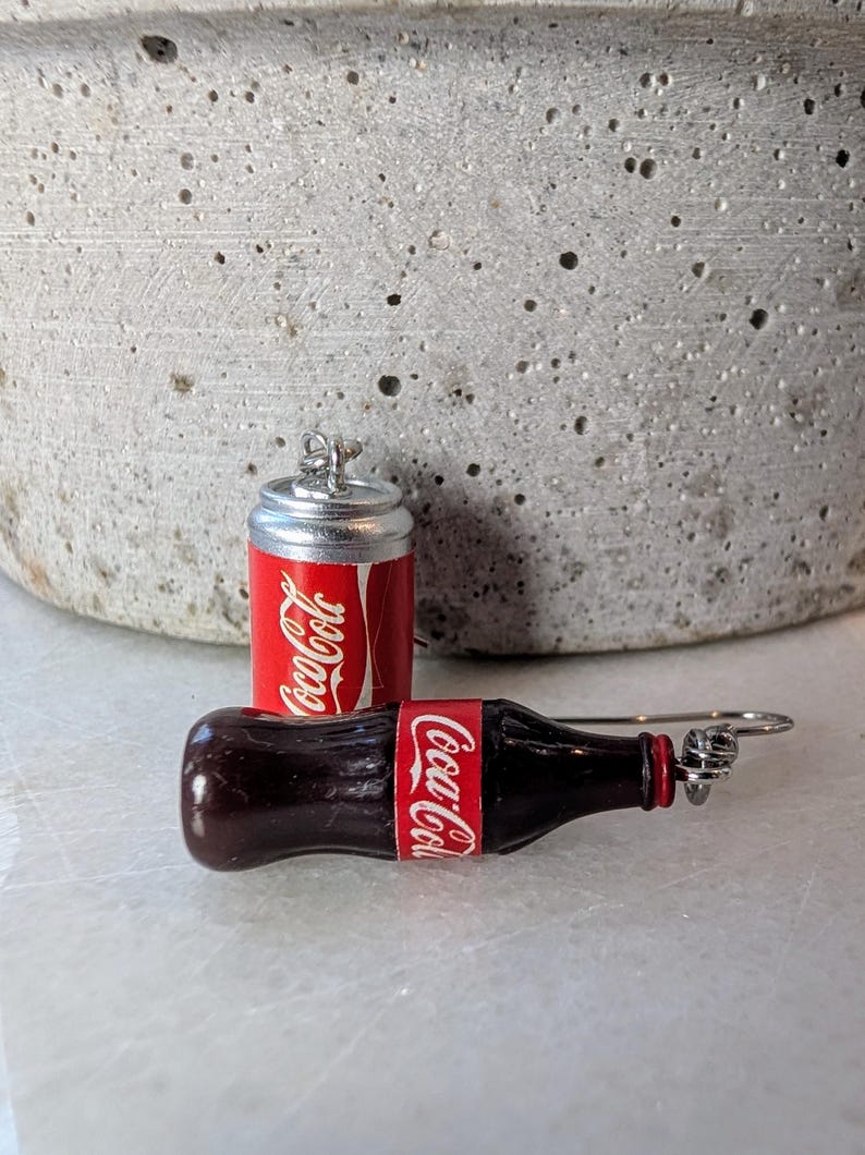 Puede incluir: Un par de pendientes con tem&aacute;tica de Coca-Cola. Un pendiente es una lata de Coca-Cola en miniatura y el otro es una botella de Coca-Cola en miniatura.