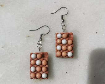 May include: A pair of earrings shaped like a carton of eggs. The carton is brown and has white eggs inside. The earrings are made of polymer clay and have silver hooks.