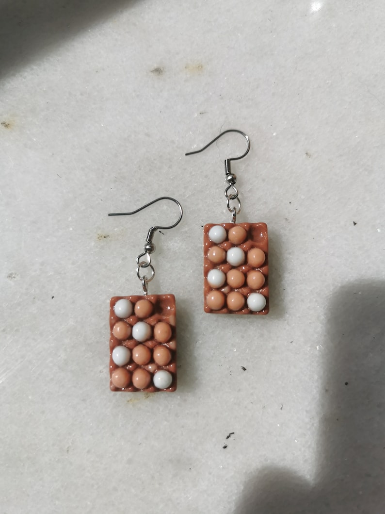 Peut inclure: Une paire de boucles d'oreilles en forme de bo&icirc;te &agrave; &oelig;ufs. La bo&icirc;te est brune et contient des &oelig;ufs blancs &agrave; l'int&eacute;rieur. Les boucles d'oreilles sont en p&acirc;te polym&egrave;re et ont des crochets argent&eacute;s.