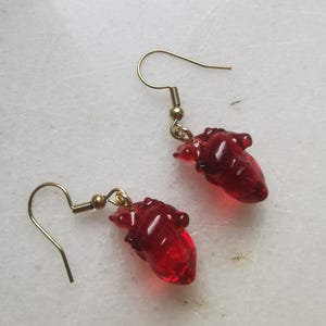 May include: A pair of gold dangle earrings with red glass heart charms. The hearts are detailed and realistic.