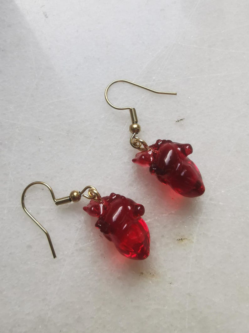 May include: A pair of gold dangle earrings with red glass heart charms. The hearts are detailed and realistic.