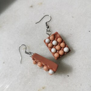 Peut inclure: Une paire de boucles d'oreilles en forme de carton d'&oelig;ufs brun et blanc. Les boucles d'oreilles sont en p&acirc;te polym&egrave;re et ont un design r&eacute;aliste de carton d'&oelig;ufs. Les boucles d'oreilles mesurent environ 2,5 cm de long et ont des crochets argent&eacute;s.