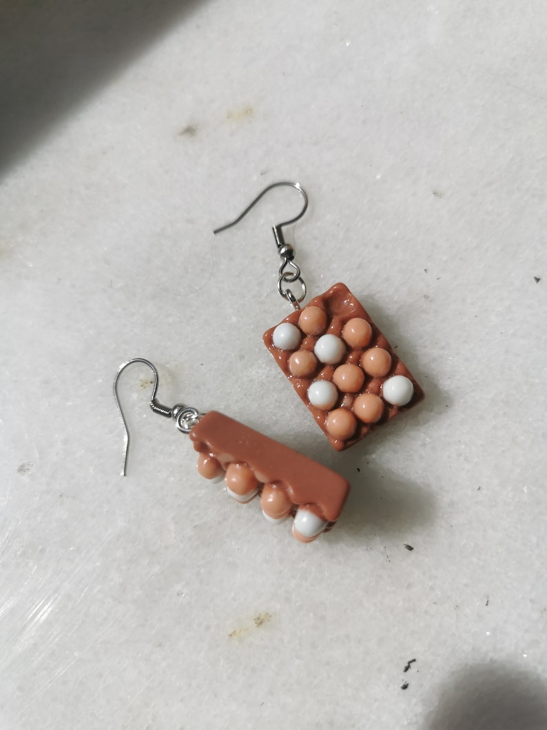 Peut inclure: Une paire de boucles d'oreilles en forme de carton d'&oelig;ufs brun et blanc. Les boucles d'oreilles sont en p&acirc;te polym&egrave;re et ont un design r&eacute;aliste de carton d'&oelig;ufs. Les boucles d'oreilles mesurent environ 2,5 cm de long et ont des crochets argent&eacute;s.