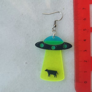 May include: A pair of neon green and black UFO earrings with a black cow silhouette inside. The earrings are made of acrylic and have silver hooks.