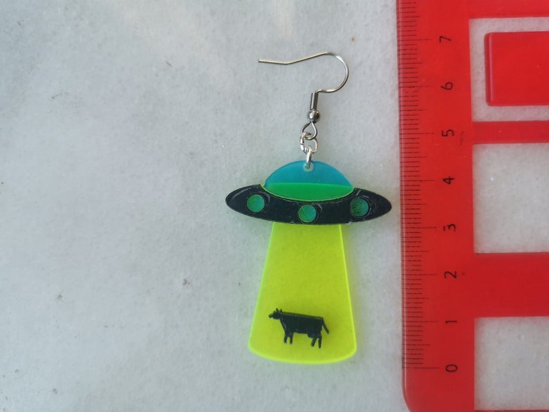May include: A pair of neon green and black UFO earrings with a black cow silhouette inside. The earrings are made of acrylic and have silver hooks.