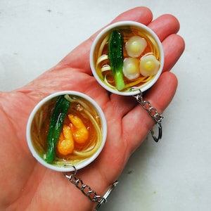 May include: Two miniature bowls of ramen noodle soup on keychains. One bowl has shrimp and green onions, the other has dumplings and green onions.