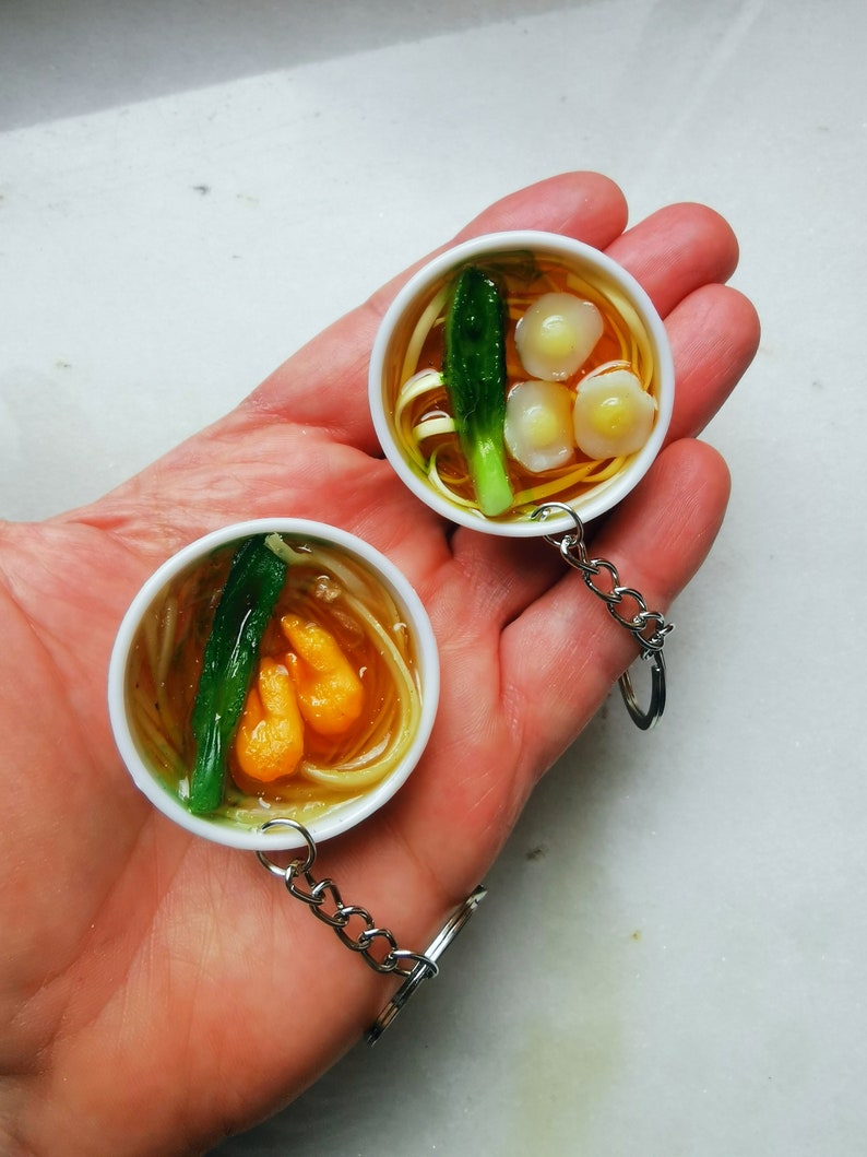 May include: Two miniature bowls of ramen noodle soup on keychains. One bowl has shrimp and green onions, the other has dumplings and green onions.