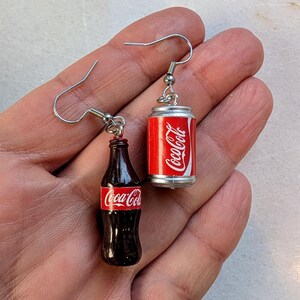 Puede incluir: Un par de pendientes colgantes de plata con abalorios en miniatura de botella y lata de Coca-Cola. La botella es marr&oacute;n con una etiqueta de Coca-Cola roja. La lata es plateada con una etiqueta de Coca-Cola roja.