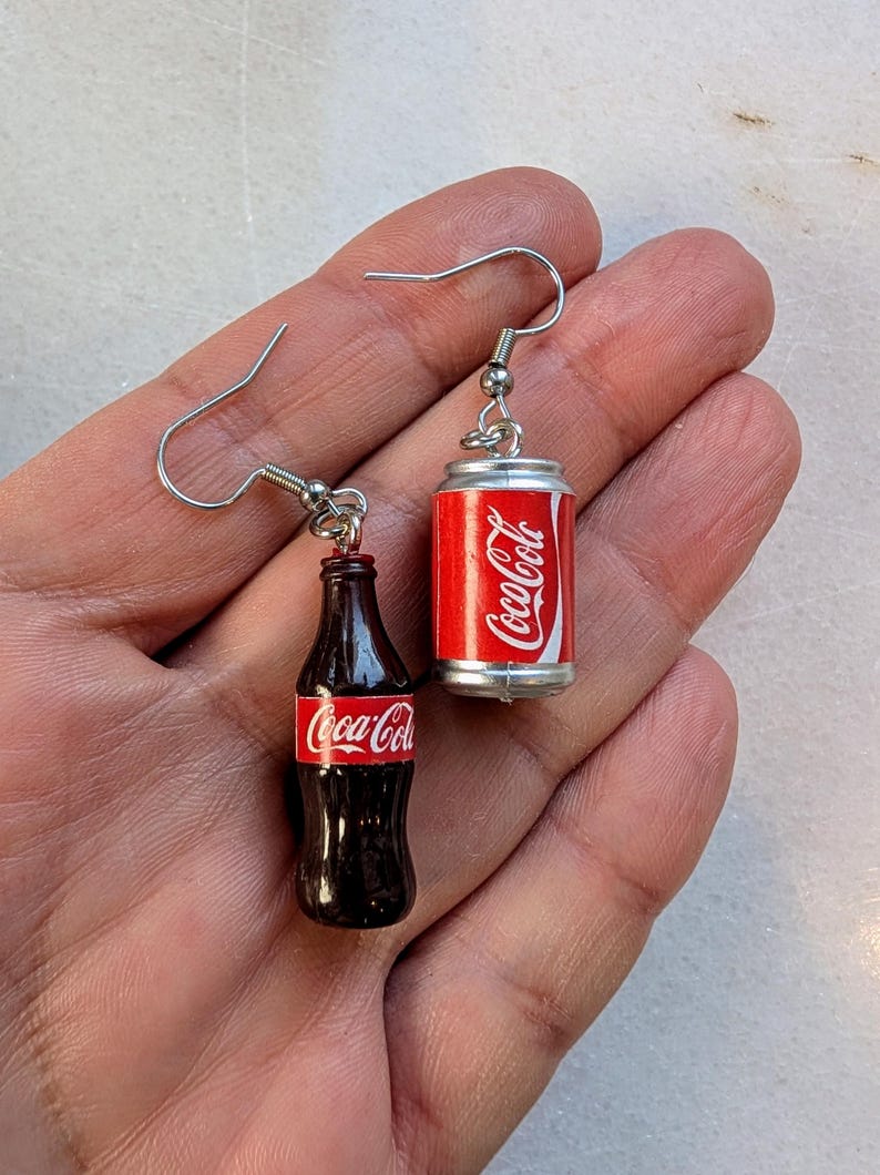 Puede incluir: Un par de pendientes colgantes de plata con abalorios en miniatura de botella y lata de Coca-Cola. La botella es marr&oacute;n con una etiqueta de Coca-Cola roja. La lata es plateada con una etiqueta de Coca-Cola roja.