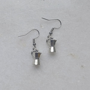 May include: A pair of silver-toned dangle earrings featuring miniature moka pot charms. Each earring is suspended from a simple metal hook wire, showcasing the detailed shape of a traditional stovetop espresso maker in a metallic finish.