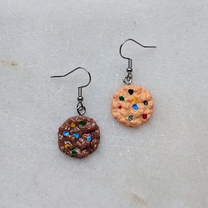 Puede incluir: Un par de pendientes colgantes con dijes circulares en forma de galleta. Un dije es de color marrón oscuro con chispas de colores y el otro es de color beige claro con chispas de colores. Ambos cuelgan de ganchos de metal plateado.