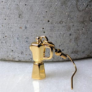 May include: A gold-colored earring shaped like a coffee maker. The earring has a small handle and a curved hook. The background is a blurred gray and white surface.