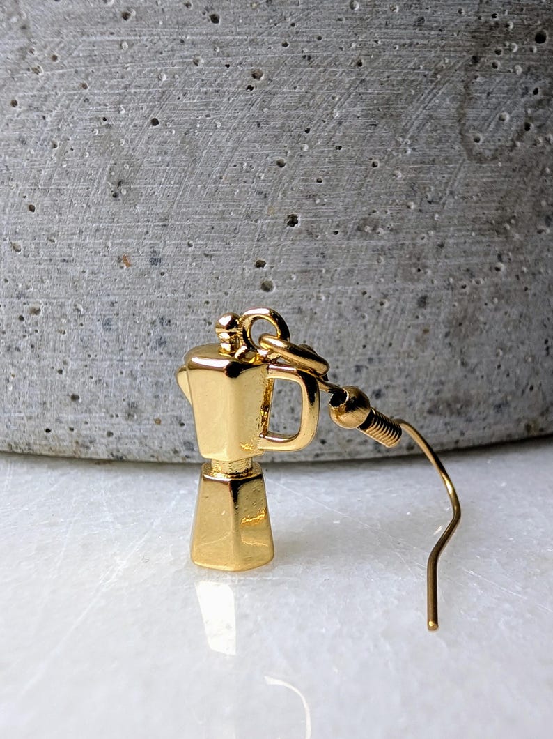 May include: A gold-colored earring shaped like a coffee maker. The earring has a small handle and a curved hook. The background is a blurred gray and white surface.
