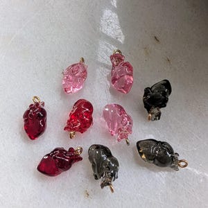 May include: Assortment of glass heart charms in various colors, including pink, red, and black. The charms have a gold loop for attaching to jewelry.