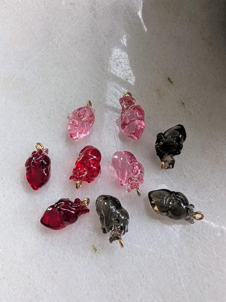 May include: Assortment of glass heart charms in various colors, including pink, red, and black. The charms have a gold loop for attaching to jewelry.