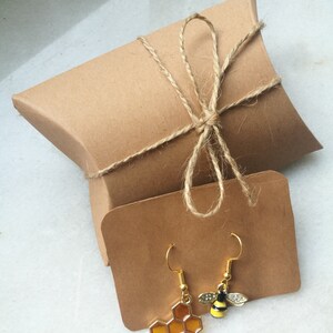 K&ouml;nnte beinhalten: Ein Paar goldfarbene Ohrringe mit einem Wabenmuster und einem Bienenanh&auml;nger. Die Ohrringe sind in einem braunen Papierumschlag verpackt, der mit Schnur gebunden ist.