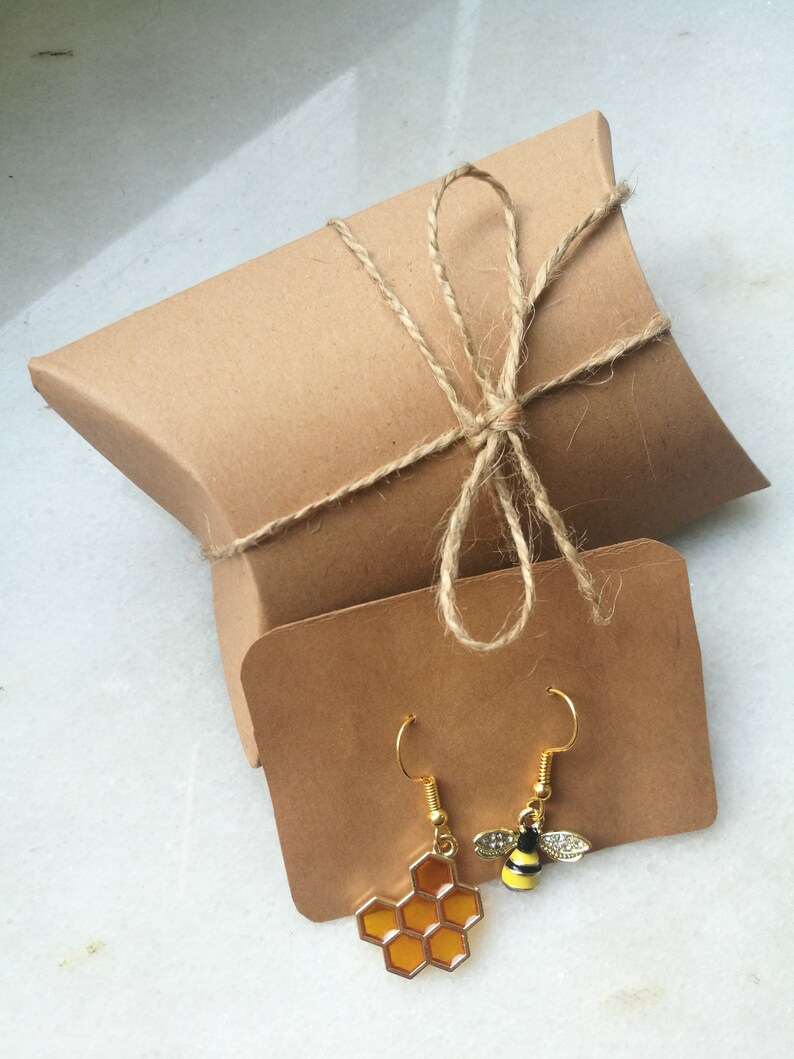 K&ouml;nnte beinhalten: Ein Paar goldfarbene Ohrringe mit einem Wabenmuster und einem Bienenanh&auml;nger. Die Ohrringe sind in einem braunen Papierumschlag verpackt, der mit Schnur gebunden ist.