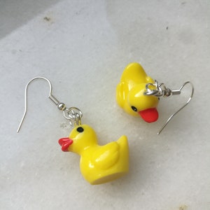Puede incluir: Un par de pendientes de pato de goma amarillo con ganchos plateados. Los patos tienen picos rojos y ojos negros.