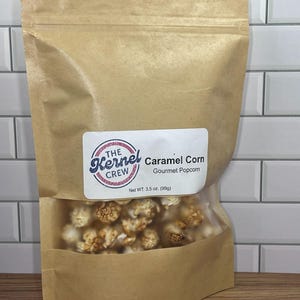 May include: A brown paper bag of caramel corn with a clear window. The label reads "The Kernel Crew Caramel Corn Gourmet Popcorn" and "Net WT. 3.5 oz. (99g)". The popcorn is a mix of golden brown and white.