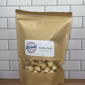 May include: A brown paper bag of kettle corn with a clear window. The bag has a label that reads "The Kernel Crew Kettle Corn Gourmet Popcorn". The net weight is 2 oz (56g). The background is a white brick wall.